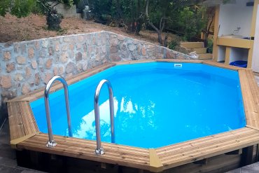 Piscina esagonale allungata in legno 5,60 m rivestimento azzurro e scaletta inox. Realizzata su piattaforma in piastrelle grigie, perfetta per momenti di svago immersi nella natura.